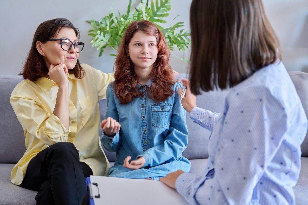 A woman and a teenage girl sit on a couch talking to another woman, who is holding a clipboard, in an indoor setting with a plant in the background.