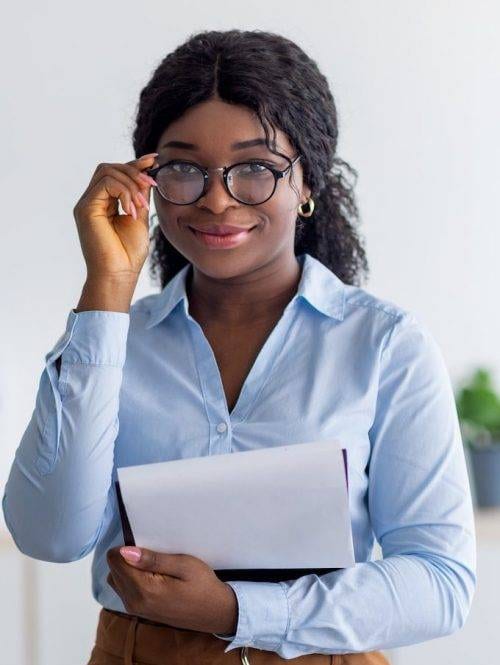 A woman wearing glasses and a light blue shirt holds a closed laptop or binder, smiling slightly while adjusting her glasses.