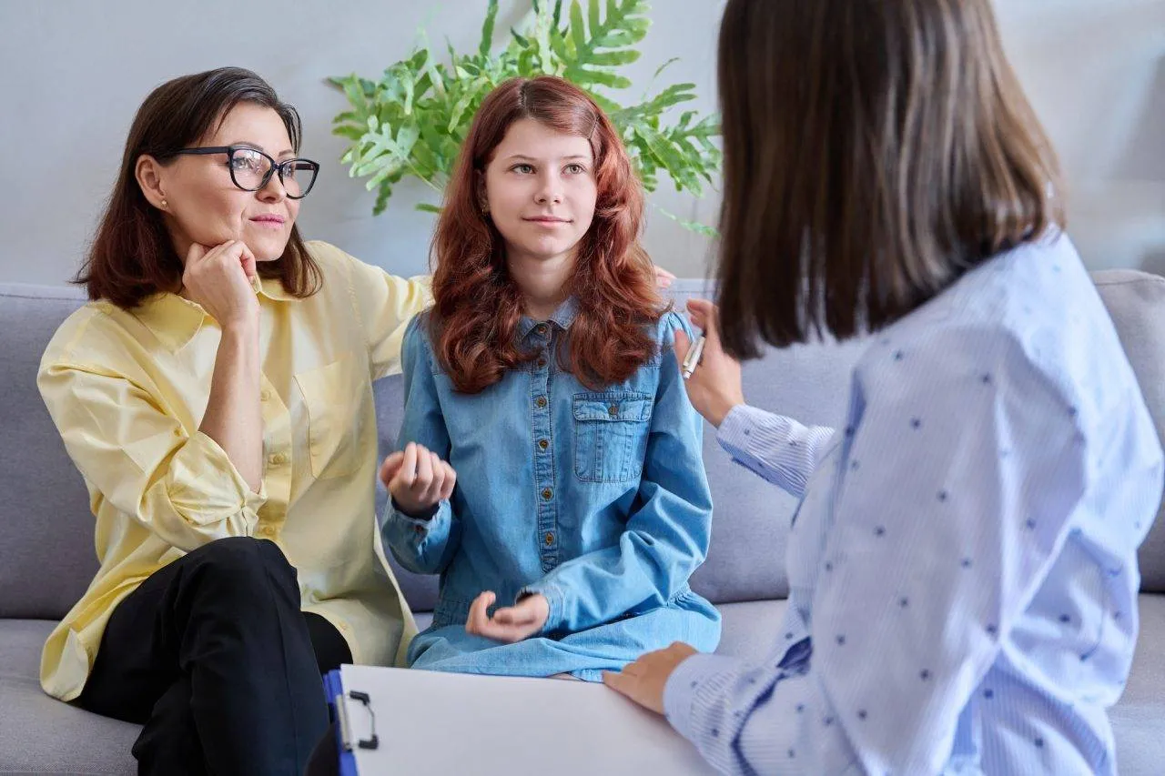 A woman and a teenage girl sit on a couch talking to another woman, who is holding a clipboard, in an indoor setting with a plant in the background.