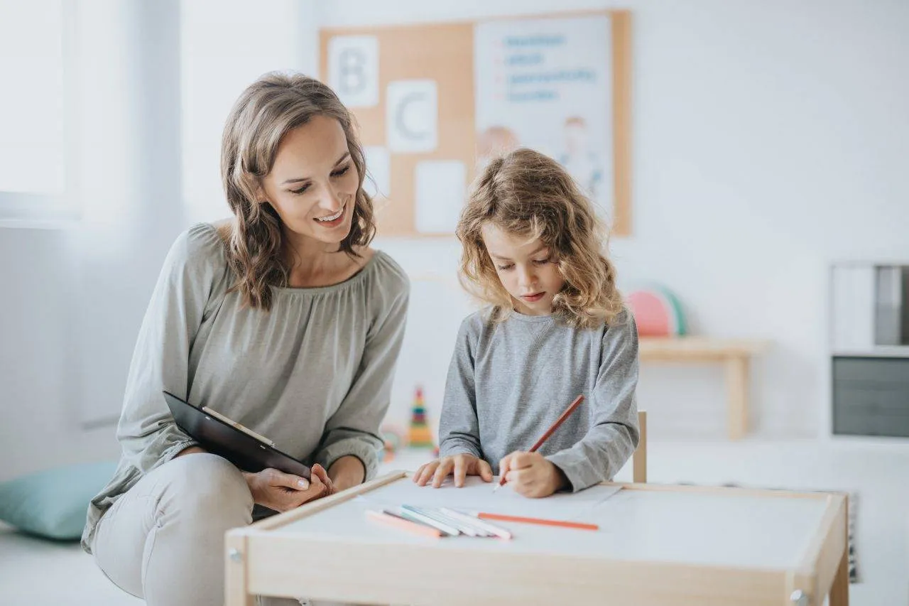 An adult sits beside a young child who is drawing with colored pencils at a table; the adult holds a clipboard and watches the child.