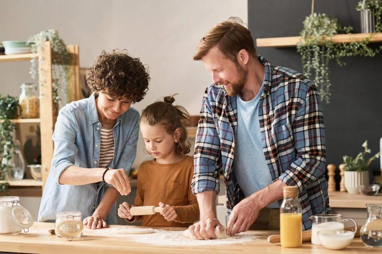 Three people, two adults and a child, are making dough together on a kitchen counter, surrounded by baking ingredients and utensils.