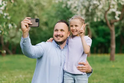Caucasian Dad and Daughter Smile and Take Selfie on Their Phone while Walking in Summer Park
