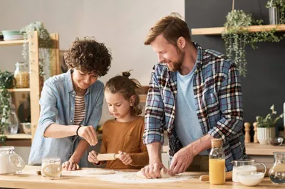 Three people, two adults and a child, are making dough together on a kitchen counter, surrounded by baking ingredients and utensils.
